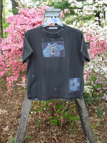 1997 Short Sleeved Tee Butterfly Ebony Size 0 on a clear hanger outdoors, showcasing stylized appliqué designs—a fish or leaf near the chest and a small flower at the lower front.
