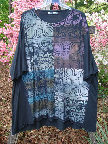 Barclay Art Tunic Tee Field Friends True Black OSFA drapes on a hanger, showcasing its oversized silhouette, dolman sleeves, and batik-like floral motifs, set against blooming azaleas outdoors.