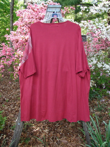 Barclay Art Tunic Tee Trailing Floral Deep Ruby OSFA, draped over a stepladder in a garden, showcases its loose fit, drop shoulders, three-quarter sleeves, and unique hemline against a floral backdrop.