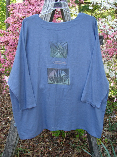 A 1995 Silk Noile Long Sleeved Top Leaf Fish Butterfly Dusk OSFA hangs on a wooden ladder, showcasing loose-cut slate-blue fabric with a stylized butterfly, fish, and geometric graphic on the front.