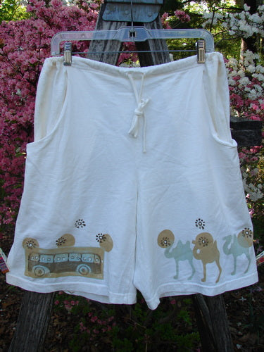 2000 Bushel Short Bus Travel Milk Size 1: White knee-length drawstring shorts with side pockets, featuring a bus and camel graphic along the hem, hanging outdoors against a backdrop of flowering shrubs.