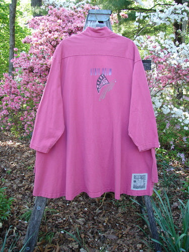 1997 Work Jacket Tiny Moon Geranium OSFA displayed on a wooden frame, showcasing its long sleeves, shirttail hemline, and intricate back graphic, set against a garden backdrop with flowering shrubs.
