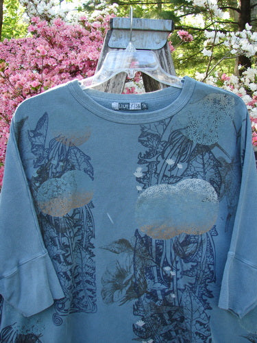 Barclay Fleece Banded Sleeve Dolman Top Moon Garden Sage Dust OSFA hangs outdoors, showcasing a faded blue-gray tunic with floral print, dolman sleeves, and a LIVE FISH tag, surrounded by vibrant flowering shrubs.