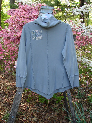 Barclay Patched Thermal Cowl Neck Roo Tunic Gray Day Size 1 displayed on a wooden frame, showcasing its cowl neck, kangaroo pocket, exposed seams, and asymmetrical hem against a floral backdrop.