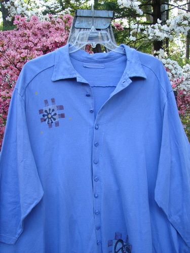 1997 Work Jacket Water Can Skylark OSFA: A light-blue long-sleeved blouse with button-front, featuring geometric motifs on chest, hanging on a clear hanger against a backdrop of flowering trees.