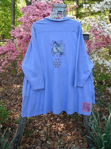 1997 Work Jacket Water Can Skylark OSFA, a loose-fitting, long-sleeved lavender top with overlapping circular graphic on the back, hangs outdoors on a rustic stand amidst blooming azaleas.