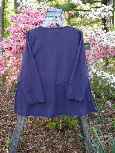 Barclay Long Sleeved A Line Tee Celtic Journey Purple Royal Size 1, draped on a wooden trellis outdoors, showcasing a rounded neckline, swinging A-line shape, and long sleeves amidst blooming shrubs.