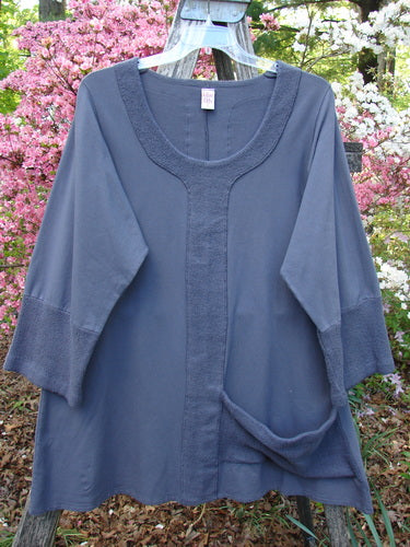 Barclay Cotton Lycra Celtic Moss Single Pocket Tunic Unpainted Grey Plum Size 1, features a scoop neckline, three-quarter sleeves, front pocket, and textured trim, hanging outdoors with flowering shrubs in the background.