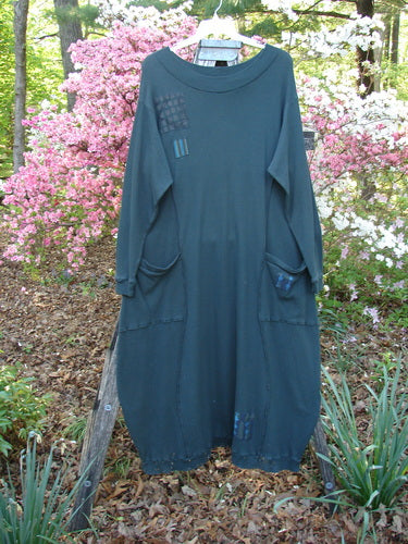 Barclay Patched Thermal Curve Dress Forest Size 2, featuring a loose tunic style with a figure-eight shape, two front pockets, and small patchwork appliqués, hanging outdoors against flowering shrubs.