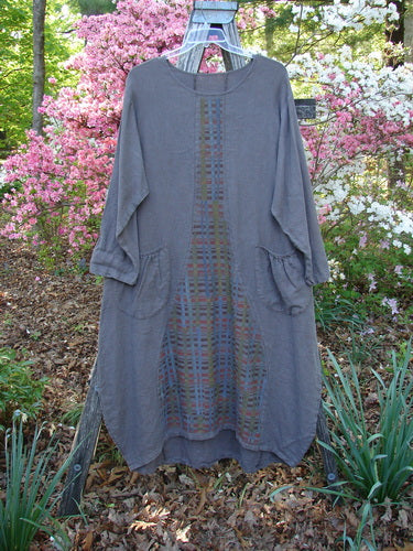 Barclay Linen Tea Ceremony Thostle Dress Cross Grid Dark Stone Size 1, a loose-fitting, soft gray tunic with multicolored plaid panel, rounded neckline, three-quarter sleeves, and large hip pockets, displayed on a wooden stand.