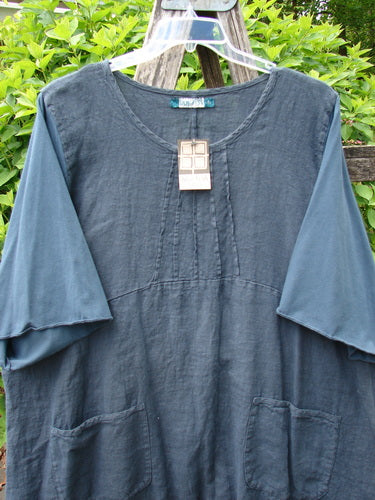 Barclay NWT Linen Cotton Sleeve Milkmaid Dress in Blue Cypress, featuring a scoop neckline, pintuck detailing, elbow-length layered sleeves, and diagonal front patch pockets, hanging outdoors on a clear plastic hanger.