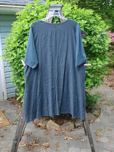 Barclay NWT Linen Cotton Sleeve Milkmaid Dress in Blue Cypress hangs on a wooden A-frame, showcasing its rounded neckline, sunrise pleats, empire waist, and diagonal pockets in an outdoor garden setting.