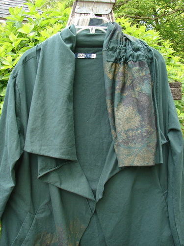 Barclay Cotton Lycra Gather Collar Open Front Jacket drapes on a hanger outdoors, showcasing its elegant flow and unique drawcord neckline, paired with camouflage-print pants over one shoulder.