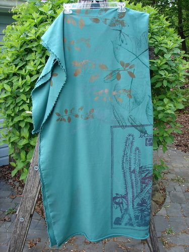 Barclay Heavy Weight Organic Sweatshirt Blanket Pony Garden Emerald OSFA draped on a wooden ladder, showcasing copper leaf motifs and teal plant scene, with raw edges, outside among leafy shrubs.