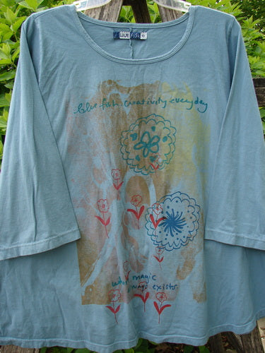 Barclay Three Quarter Sleeved A Lined Tee Creativity Everyday Dusty Sky Size 0, featuring artsy floral motifs and hand-lettered text, draped on a wooden rail, exuding a casual, bohemian charm.