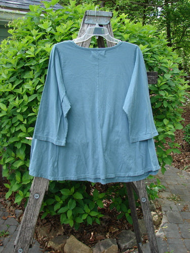 Barclay Three Quarter Sleeved A Lined Tee Creativity Everyday Dusty Sky Size 0 draped on a wooden ladder outdoors, showcasing its loose fit, rounded neckline, and curly edgings against lush greenery.