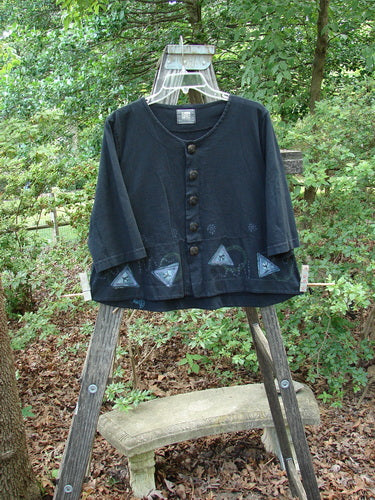 2000 4 Corners Jacket Travel Stone Black Size 1 displayed on a wooden stand, showcasing its flared shape, floral blue buttons, and distinctive swinging hem in an outdoor setting.