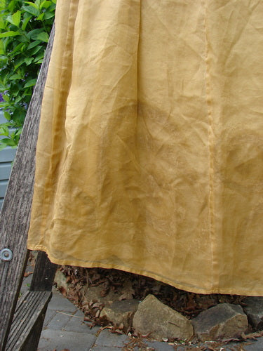 2000 Silk Organza Aios Dana Skirt Celtic Wind Bone Size 0 gracefully drapes over a wooden surface, showcasing its superior lower floating flare and soft golden exterior against a backdrop of green foliage.
