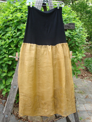 2000 Silk Organza Aios Dana Skirt Celtic Wind Bone Size 0: A two-tone skirt with a solid black upper and a shiny gold, gathered lower section, elegantly displayed against a backdrop of green shrubs.