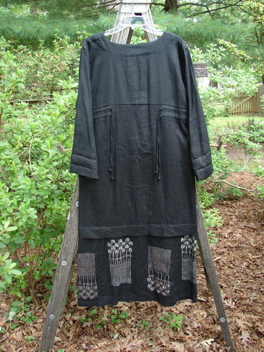 2000 Ringlet Dress Tulip Grass Black Altered Size 1 displayed on a wooden rack, showcasing its oversized pockets, sectional panels, and daisy-themed continuous hem, with slight shoulder and lower skirt alterations.