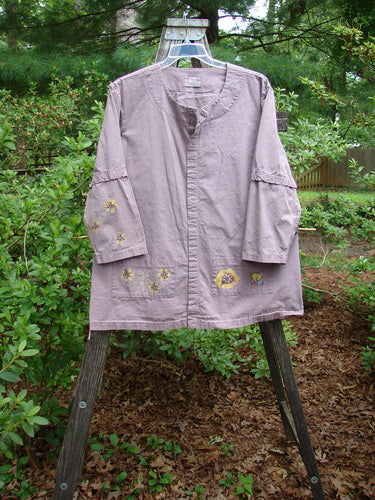 2000 Parachute Camper Jacket Florals Pale Purple Size 0 displayed on a hanger, featuring floral-themed paint, four front pockets, and snap-off sleeves, highlighting its vintage charm and unique design.