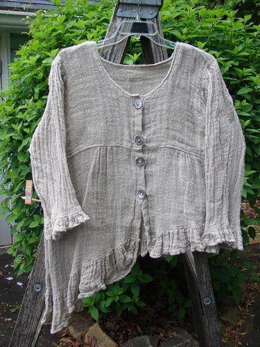Magnolia Pearl Fisherman's Weave Asymmetrical Ruffle Jacket OSFA, featuring an oatmeal-grey weave, oversized pearl buttons, three-quarter sleeves with ruffles, and an asymmetrical ruffled hem, displayed on an outdoor wooden ladder.