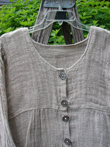 Magnolia Pearl Fisherman's Weave Asymmetrical Ruffle Jacket OSFA features oversized pearl buttons, an asymmetrical ruffled hem, and a downward curved upper bodice, hanging against rustic wood with foliage.