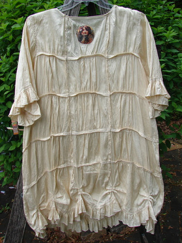 Magnolia Pearl Silk Cotton Drapery Dress Butter Creme OSFA displayed on a form. Features include elbow-length sleeves, horizontal ruffled tiers, and a decorative medallion at the neckline, set against greenery.