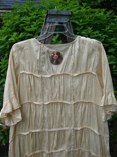 Magnolia Pearl Silk Cotton Drapery Dress Butter Creme OSFA, featuring tiered ruffles, flounced sleeves, and a vintage-style woman's portrait medallion, elegantly displayed on a wooden ladder with lush greenery backdrop.