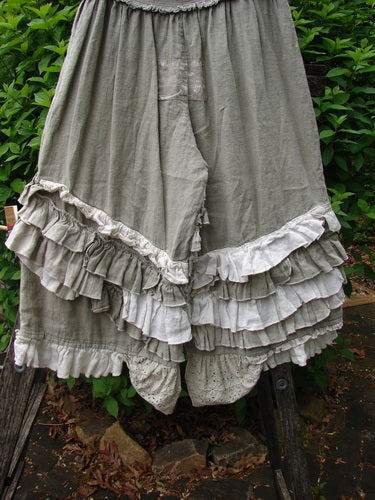 Magnolia Pearl Linen Eiffel Tower Bloomer Flaxen OSFA features muted gray-green fabric, multi-tiered ruffles, and a full smocked waistline, displayed over a wooden structure against a hedge backdrop.