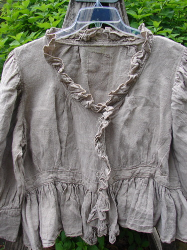 Magnolia Pearl Homespun Linen Snap Jacket Flaxen OSFA features a ruffled trim along the deeper rounded neckline and down the front, hanging on a plastic hanger amidst a backdrop of lush greenery.