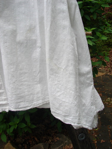 The Magnolia Pearl Cotton Voile Sweet Collar Top, a white, loosely woven garment, is draped outdoors, showcasing its angular collar, varying hem lengths, and vented sides against a natural backdrop.