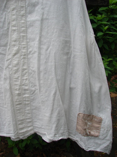 The Magnolia Pearl Cotton Voile Sweet Collar Top White OSFA, a lightweight linen garment, features a pleated panel, angular collar, and signature love patch, hanging against lush greenery.