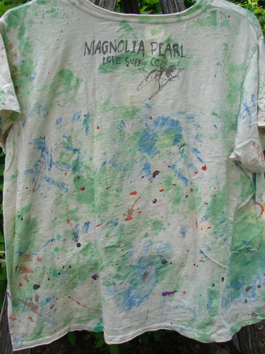 Magnolia Pearl Cotton Jersey Boyfriend Tee Songbird Floral OSFA, splattered with vibrant paint colors, features a printed MAGNOLIA PEARL LOVE Supply Co. on the back, hung outdoors, showcasing its artistic, distressed design.
