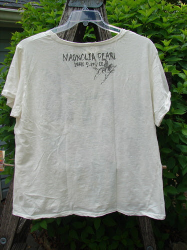 Magnolia Pearl Cotton Jersey Boyfriend Tee Iris Garden OSFA displayed on a wooden ladder, featuring off-white fabric with a rear Magnolia Pearl Love Supply Co. stamp, showcasing a super soft, drapey feel.