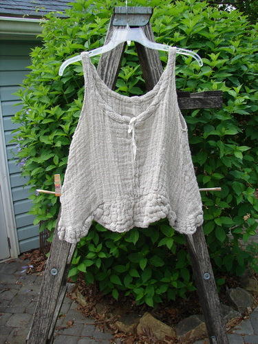 Magnolia Pearl Fisherman's Weave Ribbon Neck Top Flaxen OSFA features a light-cream, sleeveless design with tiny buttons, a tie at the bust, and a textured knit, displayed on a wooden stepladder.
