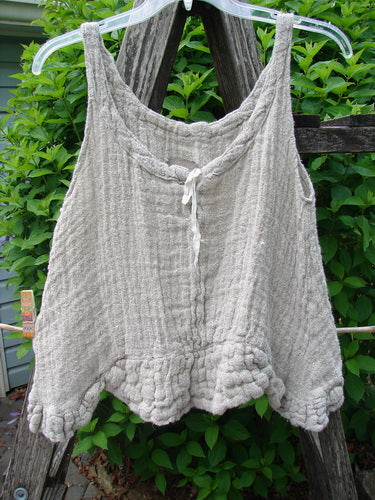 Magnolia Pearl Fisherman's Weave Ribbon Neck Top Flaxen OSFA hangs on a rustic ladder, showcasing its textured basket weave, elegant neckline tie, and scalloped hem with weighted ruffles against lush greenery.