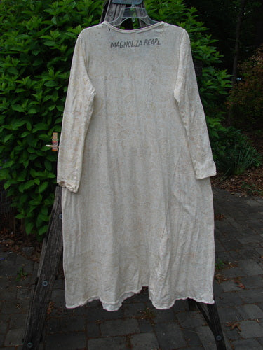 Magnolia Pearl Cotton Dylan Dress Softest Floral Naranja OSFA hangs on a wooden ladder, showcasing its vintage style with rolled neckline, tighter sleeves, and distressed details against a lush green backdrop.