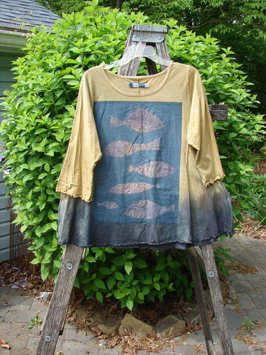 Barclay Three Quarter Sleeved Cafe Tee Sea of Fish Mottled Mustard Size 0, featuring a blue fish stencil print, hangs on a wooden stepladder, set against leafy shrubs and a patio.