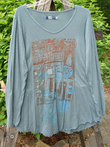 Barclay Modal Long Sleeved A Lined Tee Promo Field Dusky Sky Size 0 featuring a shallow scoop neckline, lettuce-edge hem, and earthy graphic print, hanging on a wooden stepladder amidst foliage.