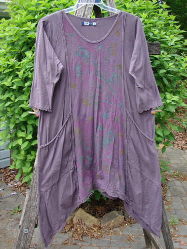 Barclay Wildflower Tunic Floral Sprig Fig Size 0, featuring asymmetrical hem, scoop neckline, elbow-length ruffled sleeves, draped pockets, and subtle front pattern, displayed on a hanger amidst greenery.