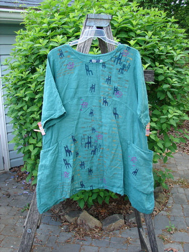 Barclay Linen Side Pocket Urchin Dress Chair Note Jade Size 0: A mid-length, teal linen dress with giraffe motifs, featuring elbow-length sleeves, rounded neckline, empire waist, and large side pockets. Displayed outdoors on a ladder.