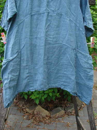 Barclay Linen Side Pocket Urchin Dress in Dusty Sky hangs outdoors, showcasing its loose fit, front patch pockets, rounded neckline, and mixed tulip garden theme, clipped with colorful clothespins.