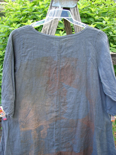 Barclay Linen Lucida Top Metallic Fern Blue Storm Size 0: A slate-gray, flowy tunic with a rounded neckline and almost three-quarter sleeves, hangs elegantly on a ladder, showcasing its lightweight linen fabric.