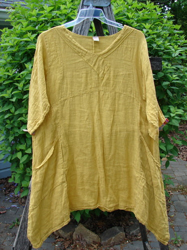 Barclay Gauze Linen Cross Over Dress Unpainted Sunshine Spice Size 1, featuring a smart V-neck, drop shoulders, empire waist seam, and side pockets, displayed against a garden backdrop.