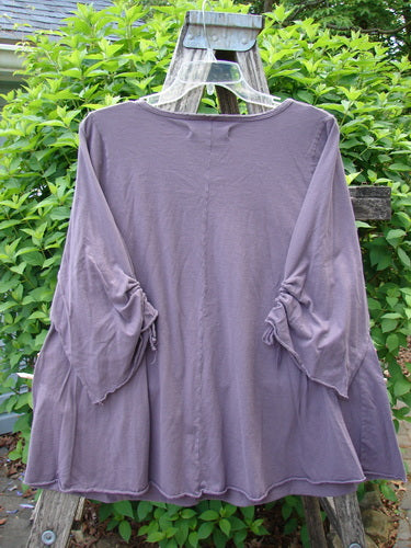 Barclay Draw Gather Sleeve Cafe Top Power Plum Size 0, showcasing loose sleeves with ties and a flared hem, hangs from a wooden ladder, highlighting its relaxed silhouette against lush greenery.