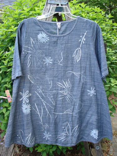 Barclay Chambray Short Sleeved Top Floral Joy Dark Denim Striation Size 0 displayed on a hanger, showcasing its slate-blue hue, hand-drawn white floral pattern, and lightweight cotton fabric against a natural backdrop.