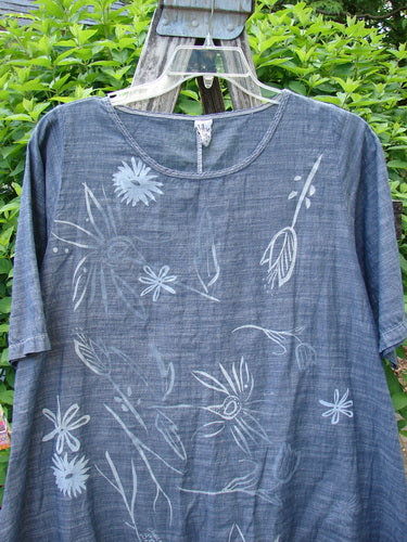 Barclay Chambray Short Sleeved Top Floral Joy Dark Denim Striation Size 0 features a relaxed, A-line shape with a scooped neckline and white botanical print, displayed on a hanger against greenery.