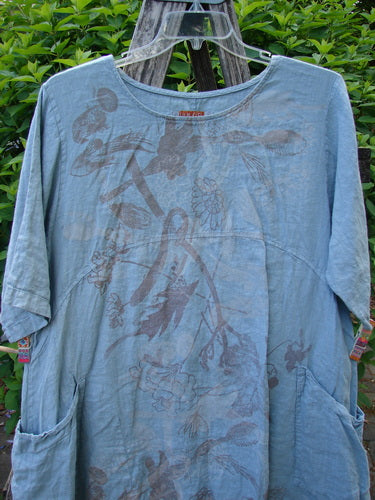 Barclay Linen Urchin Dress Leaf Floral Dusky Watercolor Size 1, showcasing a loose knee-length design with elbow sleeves, empire waist, and patch pockets, elegantly draped on a wooden ladder amidst greenery.