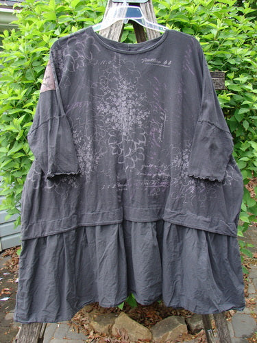 Barclay Patched Batiste Banded Flounce Esperanza Tunic Floral Storm OSFA, featuring dolman sleeves, ruffled hem, and subtle floral pattern, hangs against a leafy hedge backdrop.